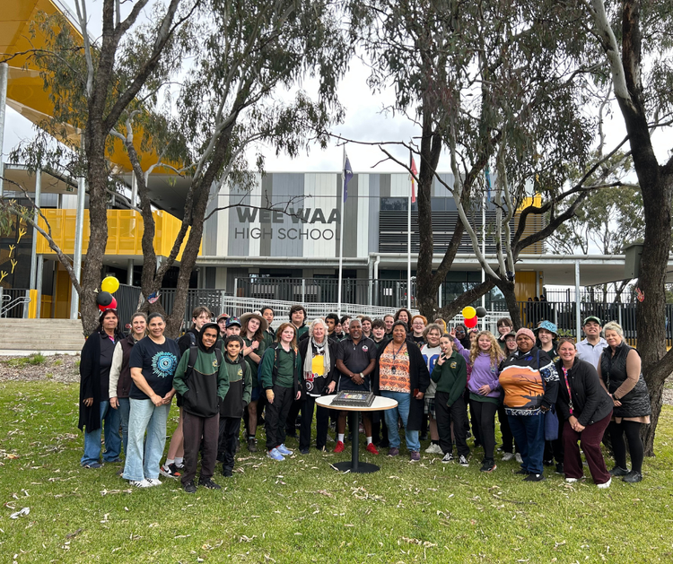 Photograph of Wee Waa High School students, staff, and community standing on the grass outside Wee Waa High School building surrounded by trees with a cake celebrating NAIDOC Week 2025