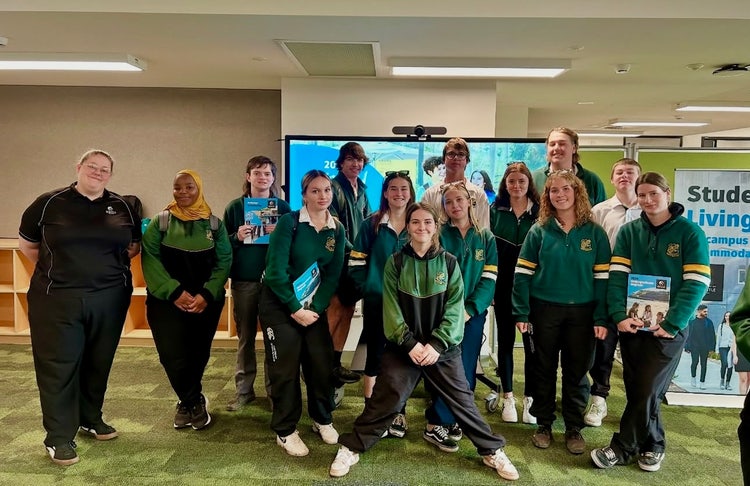 Photograph of students in Wee Waa High School uniform standing inside school library smiling with University of Newcastle staff member