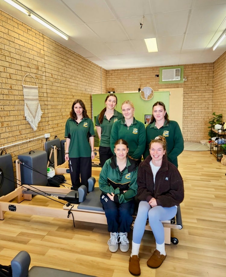 Photograph of students dressed in Wee Waa High School uniform standing inside pilates studio with pilates reformer machine and pilates instructor dressed in activewear