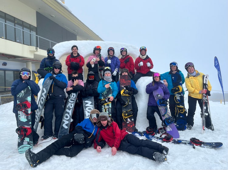 Photograph of students and staff dressed in snow gear standing outside on snow with skis and snowboards on school excursion