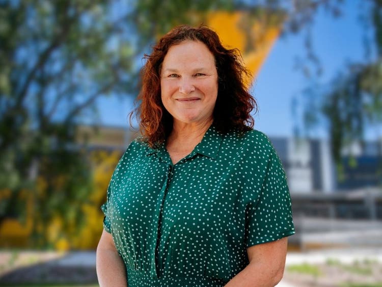 Photograph of woman smiling in front of Wee Waa High School building surrounded by trees with dark hair and green dress with white polkadots