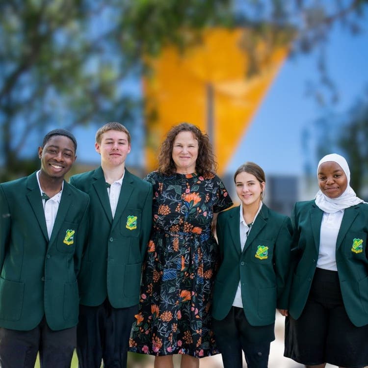 Photograph of students dressed in Wee Waa High School uniform with principal dressed in floral dress smiling in front of Wee Waa High School building surrounded by trees
