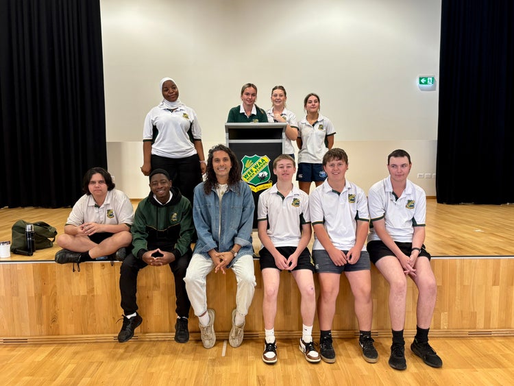 Students dressed in Wee Waa High School uniform sitting on Wee Waa High School stage inside school hall with male dressed in light pants and denim jacket at slam-poetry event